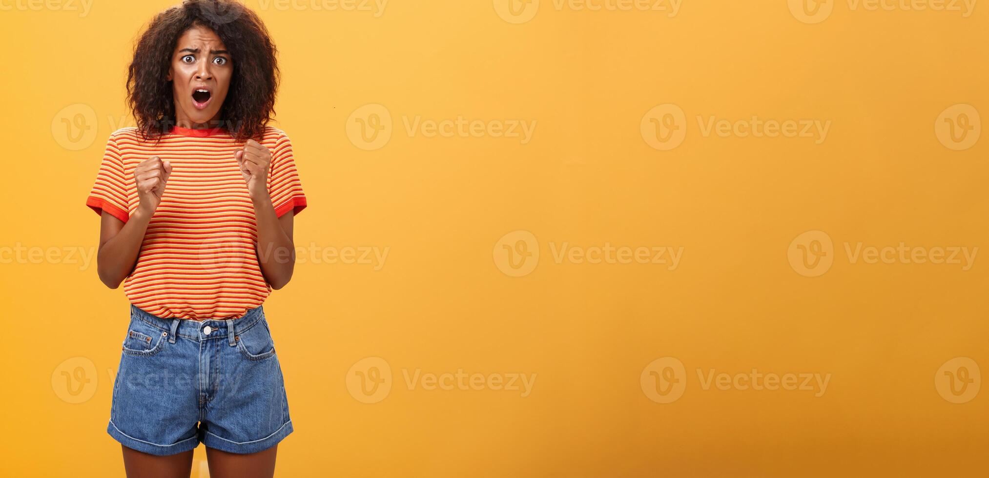 mujer acerca de a pánico escuchando impactante preocupado noticias. retrato de intenso nervioso y ansioso de piel oscura adulto niña con afro peinado apretando puños apertura boca mirando preocupado terminado naranja pared foto