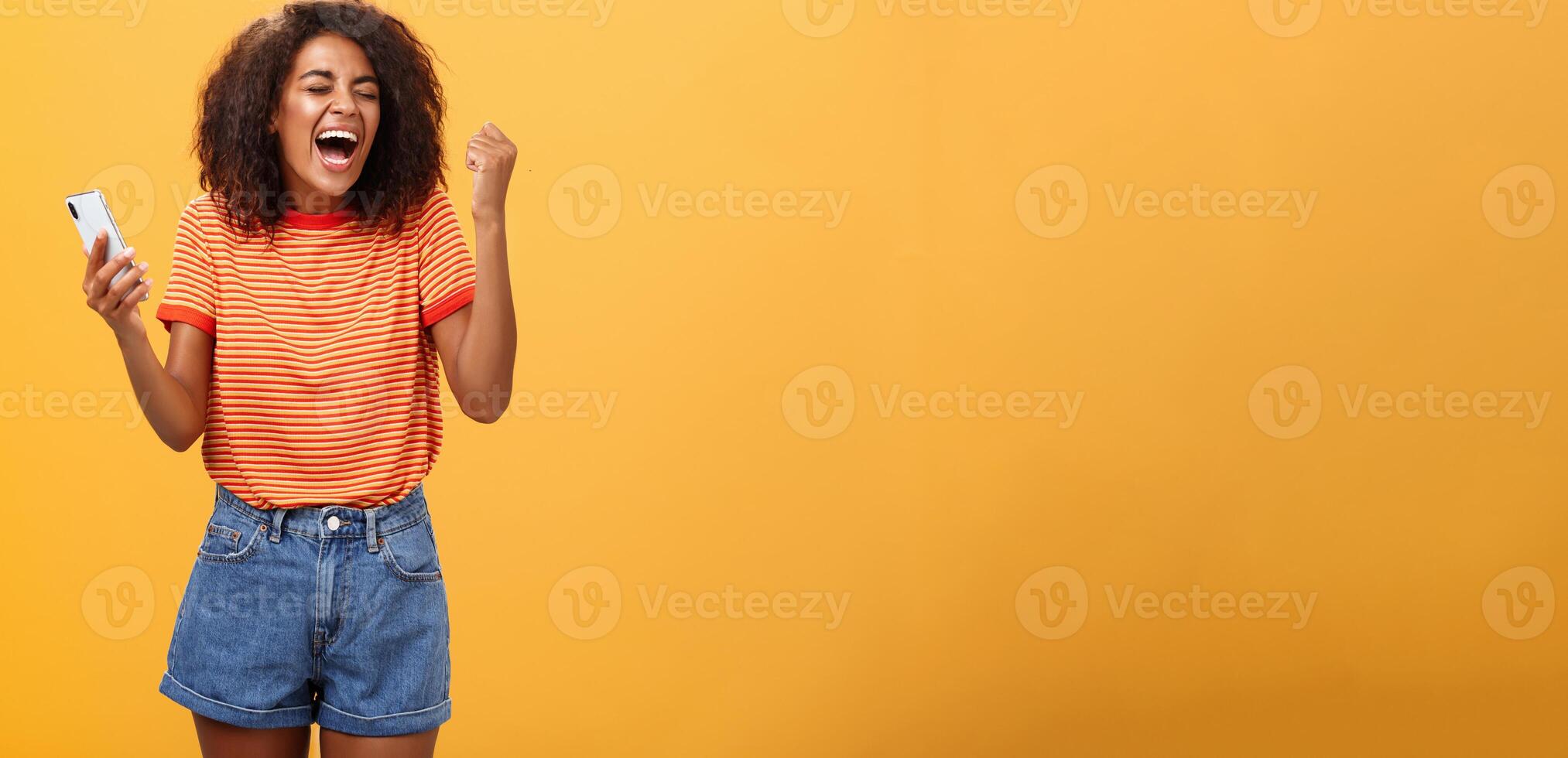 retrato de ambicioso contento joven africano americano niña Gritando desde felicidad y triunfo apretando puño en alegría y celebracion sensación emocionado y aliviado participación teléfono inteligente terminado naranja pared foto