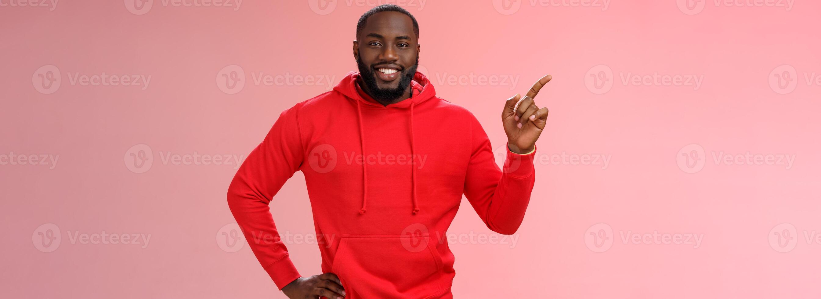 Handsome friendly-looking black guy bearded wearing casual red hoodie pointing left help customer find way directing blank space advertising product smiling happily, pink background photo