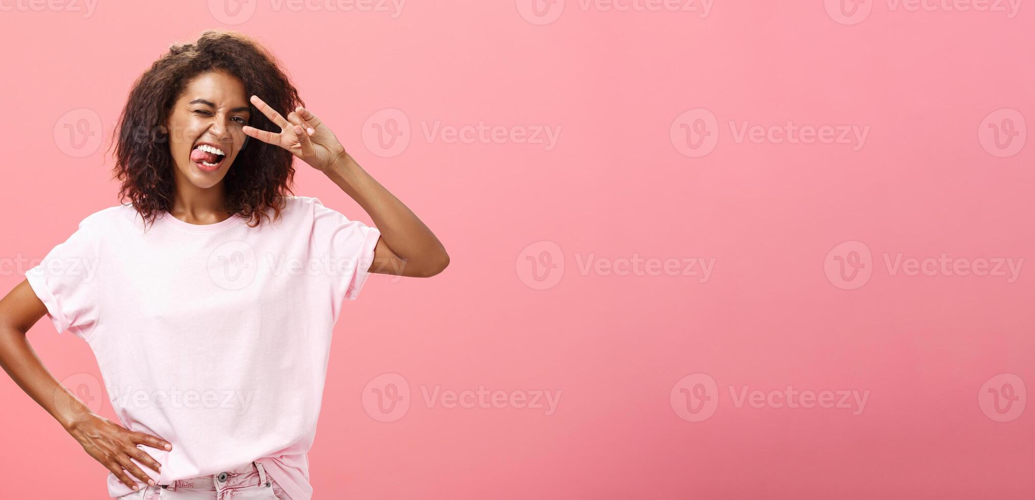 no temeroso Rápido mí mismo. alegre carismático africano americano mujer en camiseta con afro Corte de pelo demostración lengua en broma y atrevido haciendo paz firmar terminado ojo y parpadeo posando terminado rosado antecedentes foto