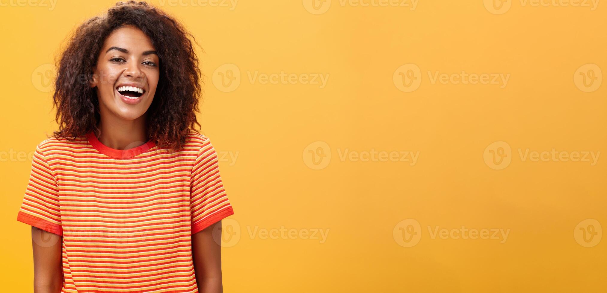 Waist-up shot of outgoing happy charming dark-skinned female with curly hairstyle laughing joyfully posing in striped trendy t-shirt over orange background enjoying nice casual conversation photo