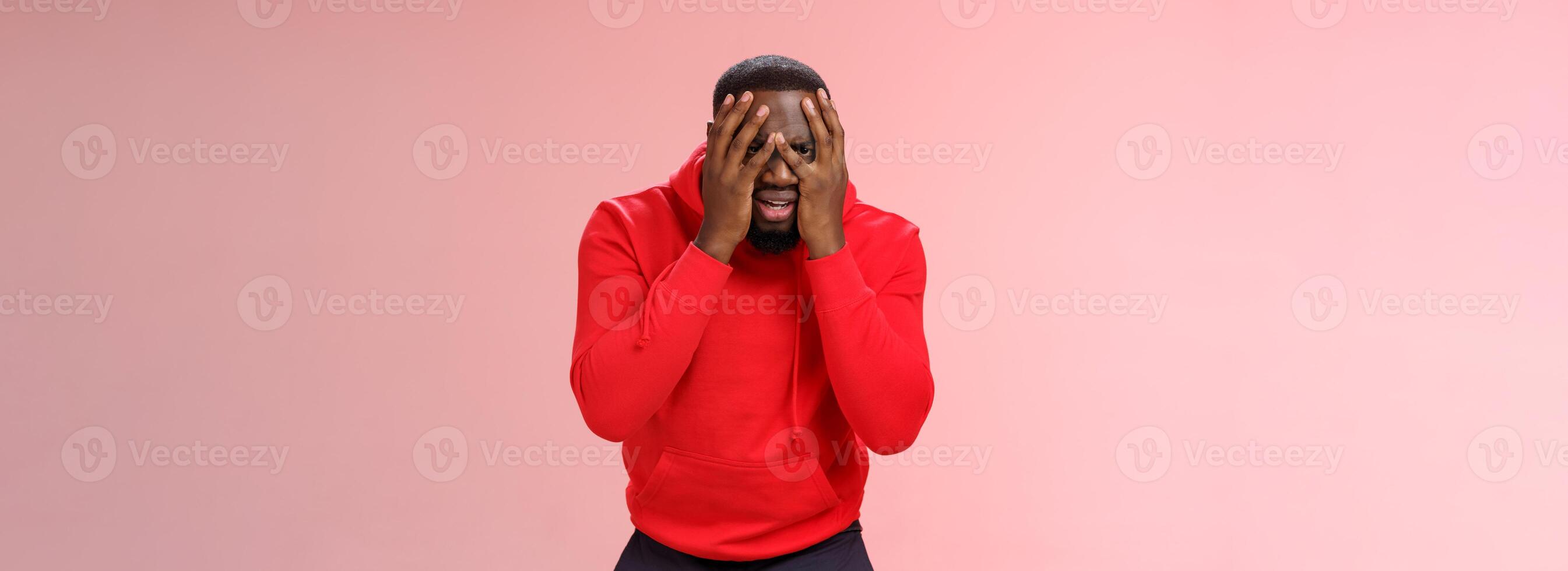Shocked upset young african american bearded guy in red hoodie facepalm hide face hands peeking through fingers sad devastated lose hope, standing sad pink background grieving feel sorrow photo