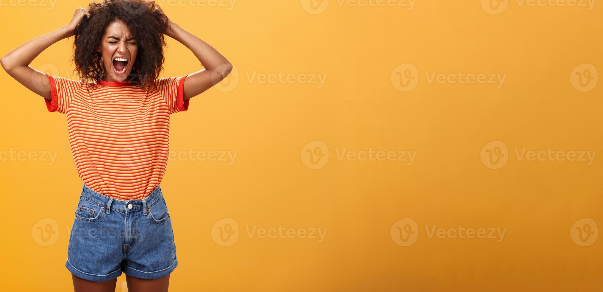 I hate life being cruel. Depressed african woman in rage and fury pulling hair out of head yelling and screaming with closed eyes losing temper having bad day hating everything over orange wall photo