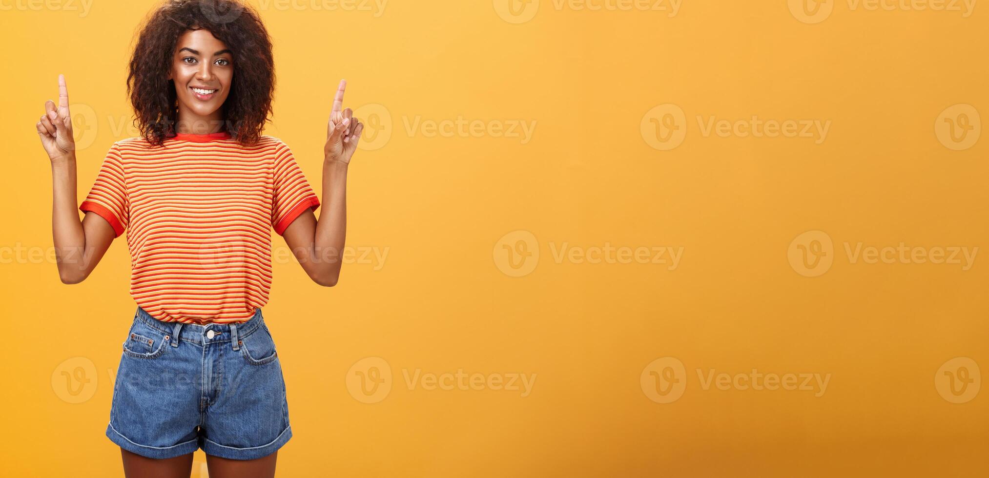 mirando solamente arriba y adelante. optimista ambicioso elegante de piel oscura hembra estudiante en a rayas frio camiseta y pantalones cortos levantamiento manos señalando hacia arriba y sonriente simpático terminado naranja pared foto