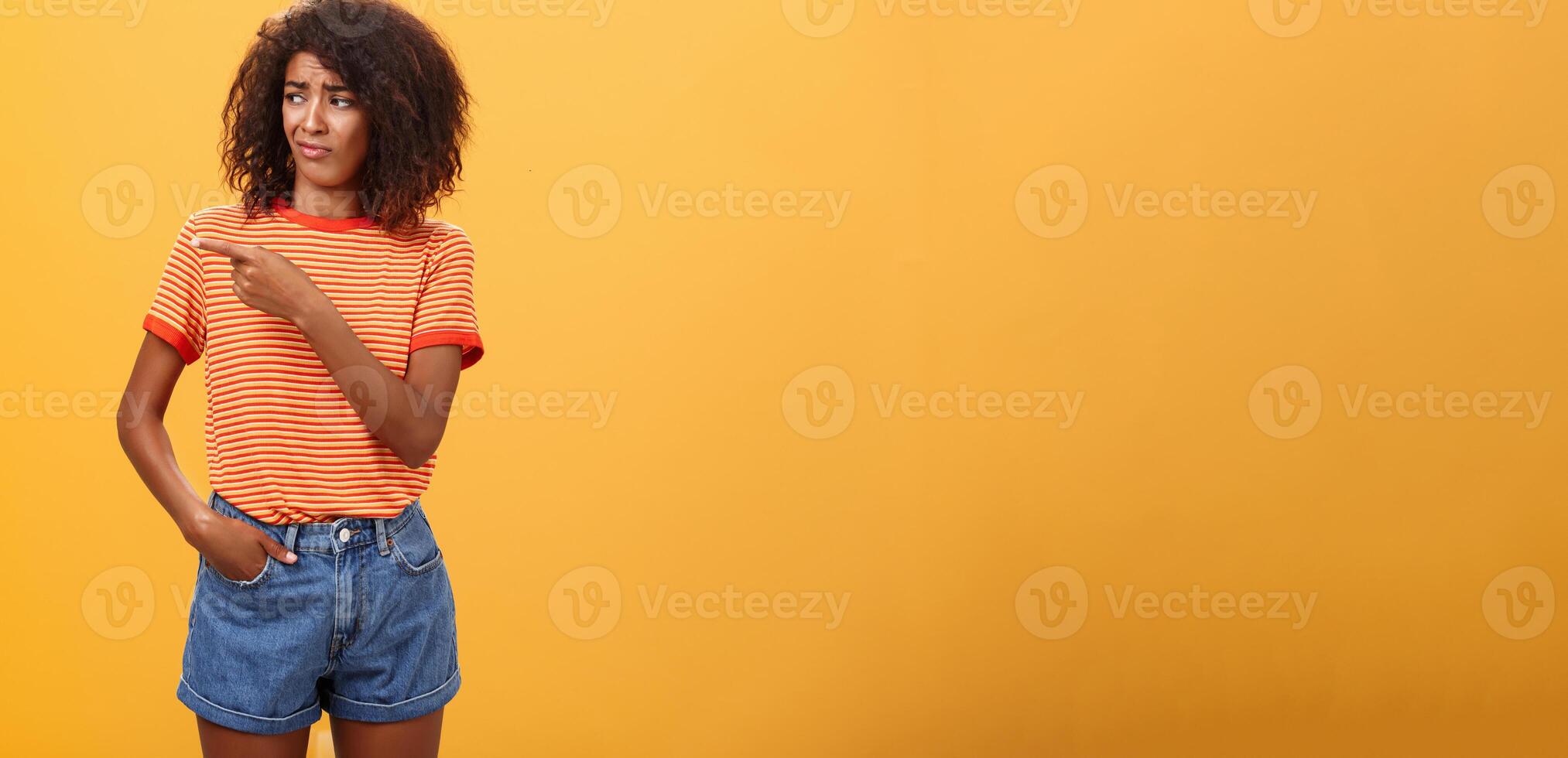 Girl looking at doubtful unimpressive perfomance of model. Portrait of displeased confused good-looking dark-skinned female with afro hairstyle looking and pointing left with scorn and indifference photo