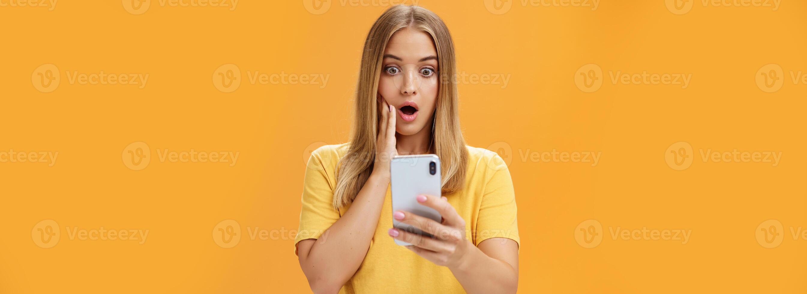 Waist-up shot of shocked and stunned emotive young woman reacting to shocking terrible news reading online from internet gasping opening mouth nervously touchin cheek staring at smartphone screen photo