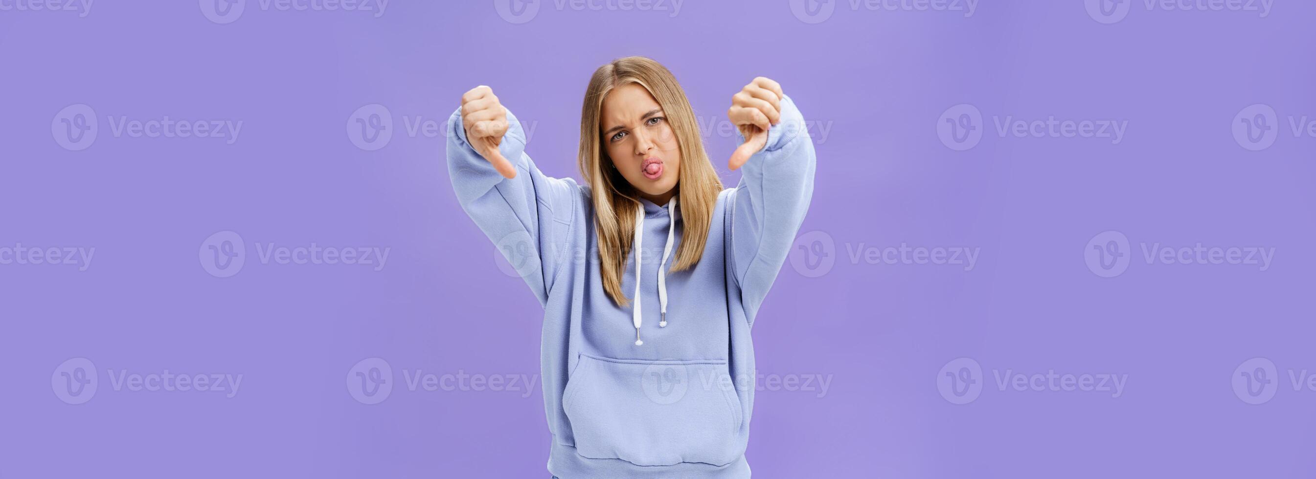 Indoor shot of moodie displeased and unimpressed cool modern female with blond hair and tanned skin tilting head sticking out tongue showing thumbs down in dissatisfaction posing against purple wall photo