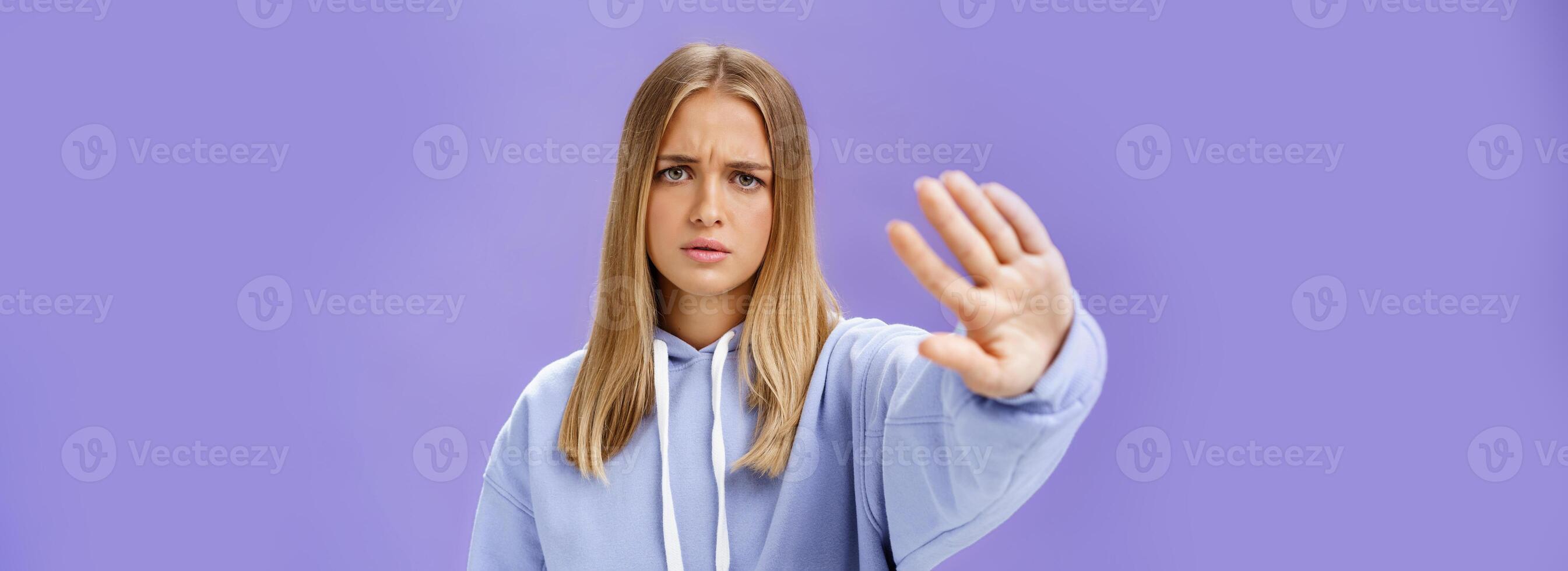 Stop photographing me. Concerned displeased young woman demanding end shooting pulling hand towards camera in no and rejection gesture frowning looking serious and displeased over blue wall photo