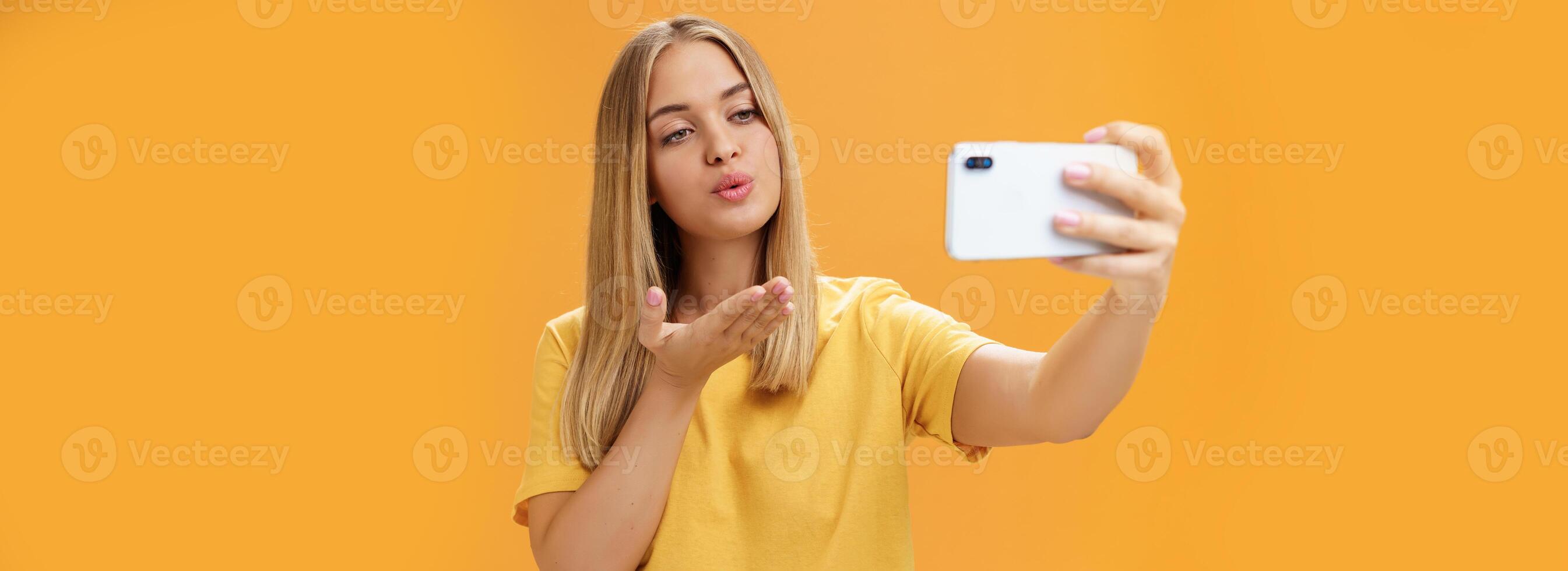 elegante glamoroso hembra Moda blogger finalizando grabación vídeo vía teléfono inteligente por enviando aire Beso a cámara, tomando selfie con sensual y confidente mirada a pantalla posando terminado naranja pared foto