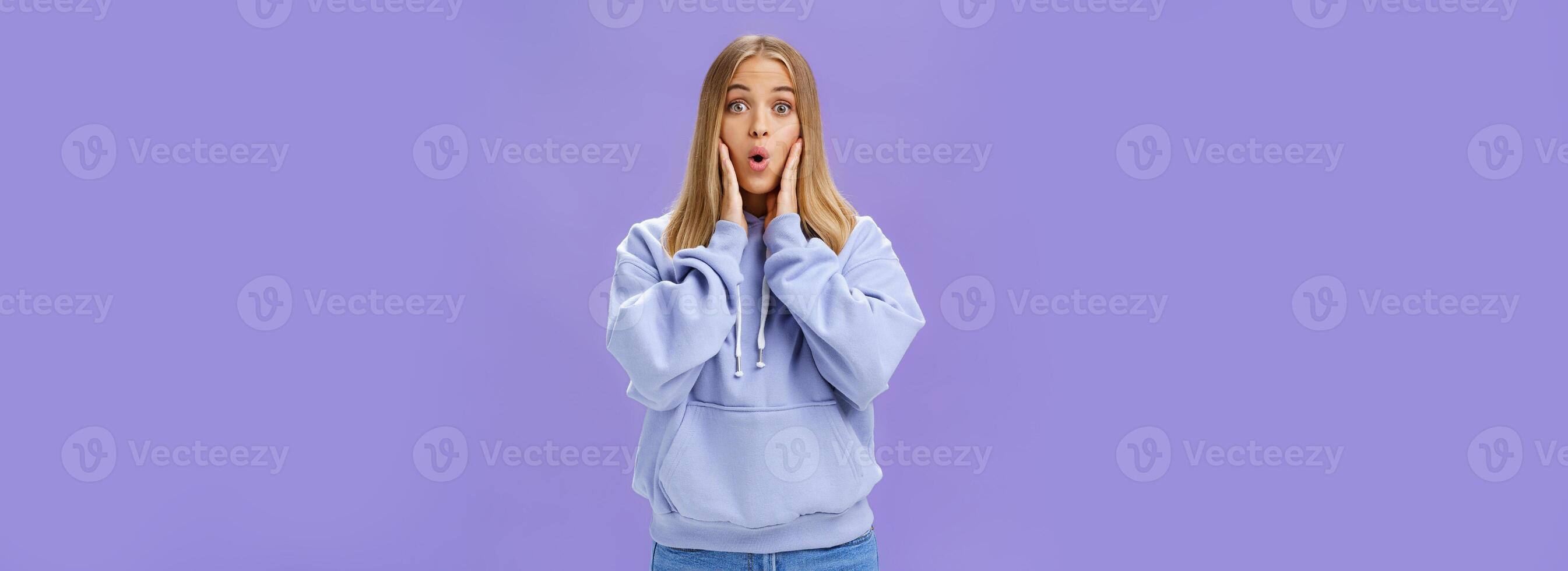 Indoor shot of amused and thrilled surprised good-looking young woman in stylish hoodie folding lips in wow sound popping eyes at camera shocked and excited learning amazing news over purple wall photo