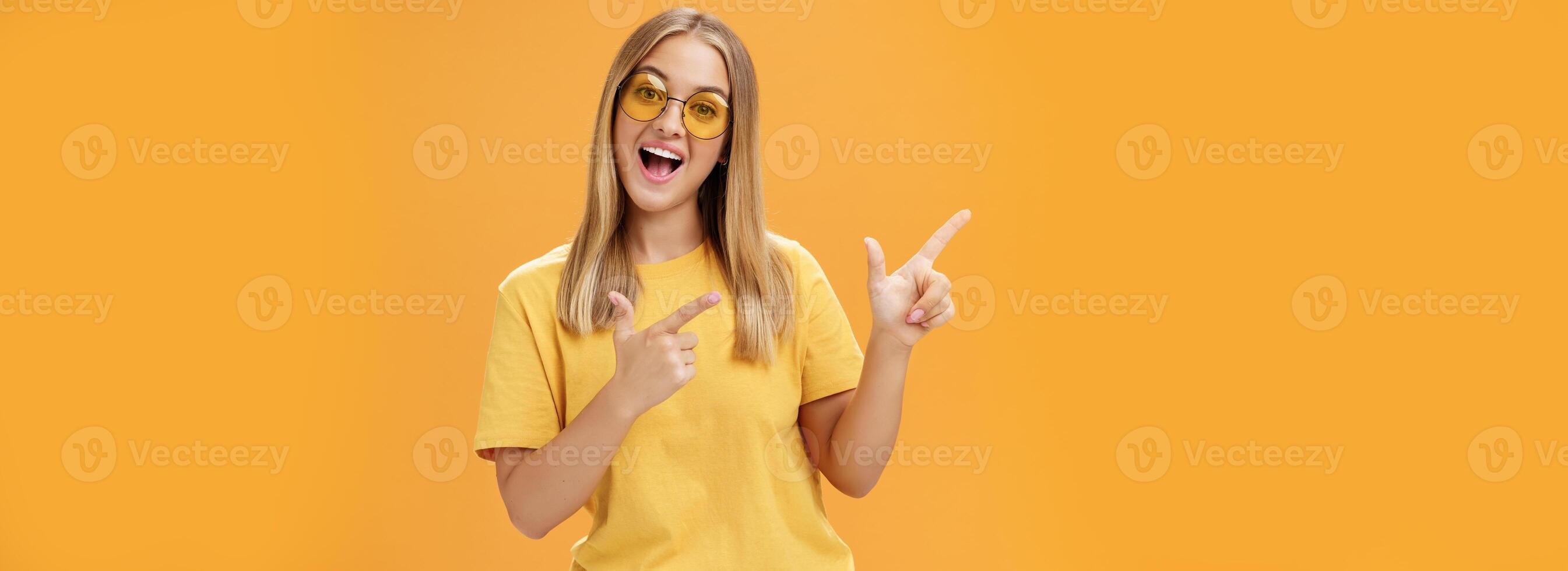 Click her watch my videos. Happy confident and outgoing charming young girl with straight hair in sunglasses and yellow t-shirt pointing at upper left corner promoting product with joyful smile photo