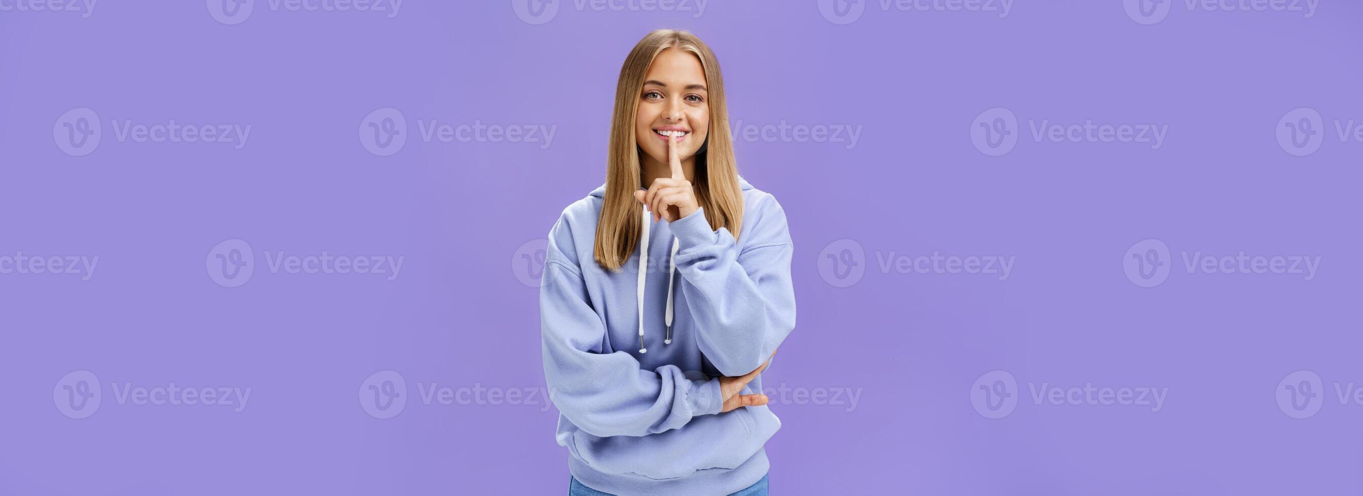 Portrait of joyful cute feminine young woman in hoodie smiling happily showing shush gesture hiding surprise asking keep secret standing amused and carefree against purple background photo