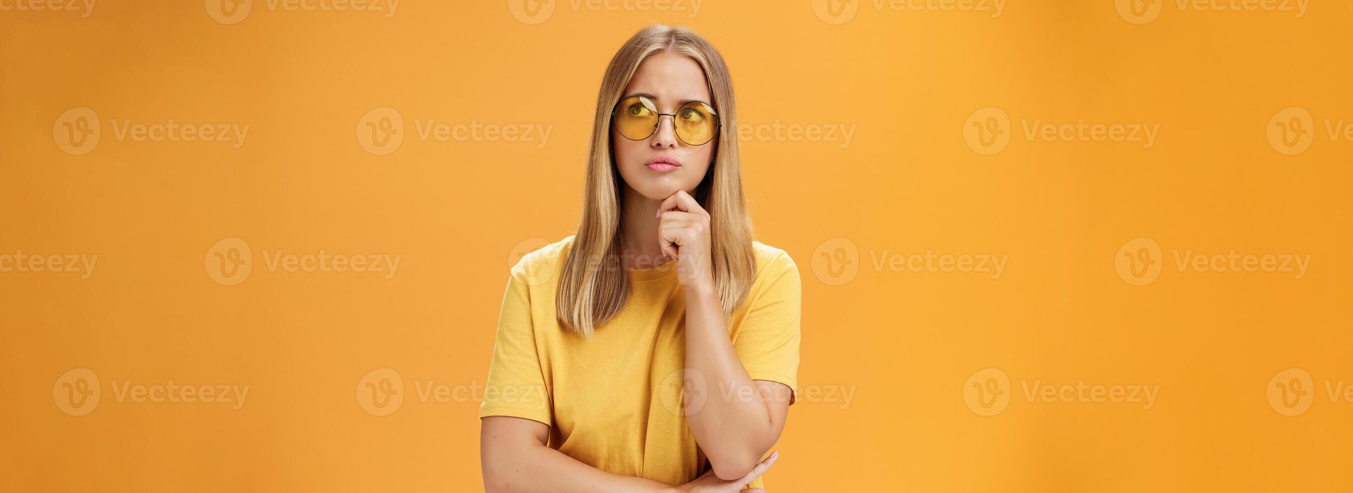 Troubled and concerned young stylish woman facing troublesome problem thinking holding hand on chin pursing lips looking at upper right corner thoughtfully standing against orange background photo