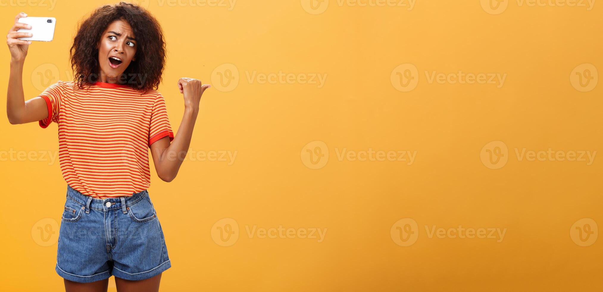 Woman recording video blog pointing at strange object behind her. Portrait of concerned and curious stylish famous internet star holding smartphone talking in cellphone camera over orange background photo