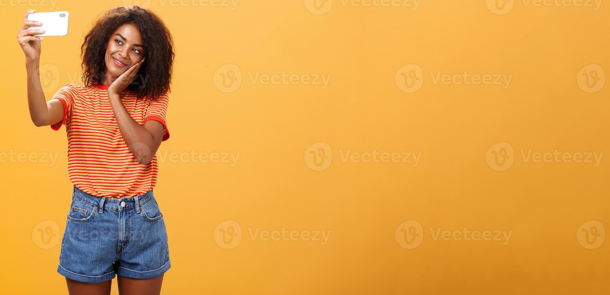 mujer hecho maquillaje todas día a enviar nuevo foto en línea tomando selfie en marca nuevo teléfono inteligente haciendo linda cara propensión cabeza en palma sonriente suavemente a dispositivo pantalla posando terminado naranja antecedentes