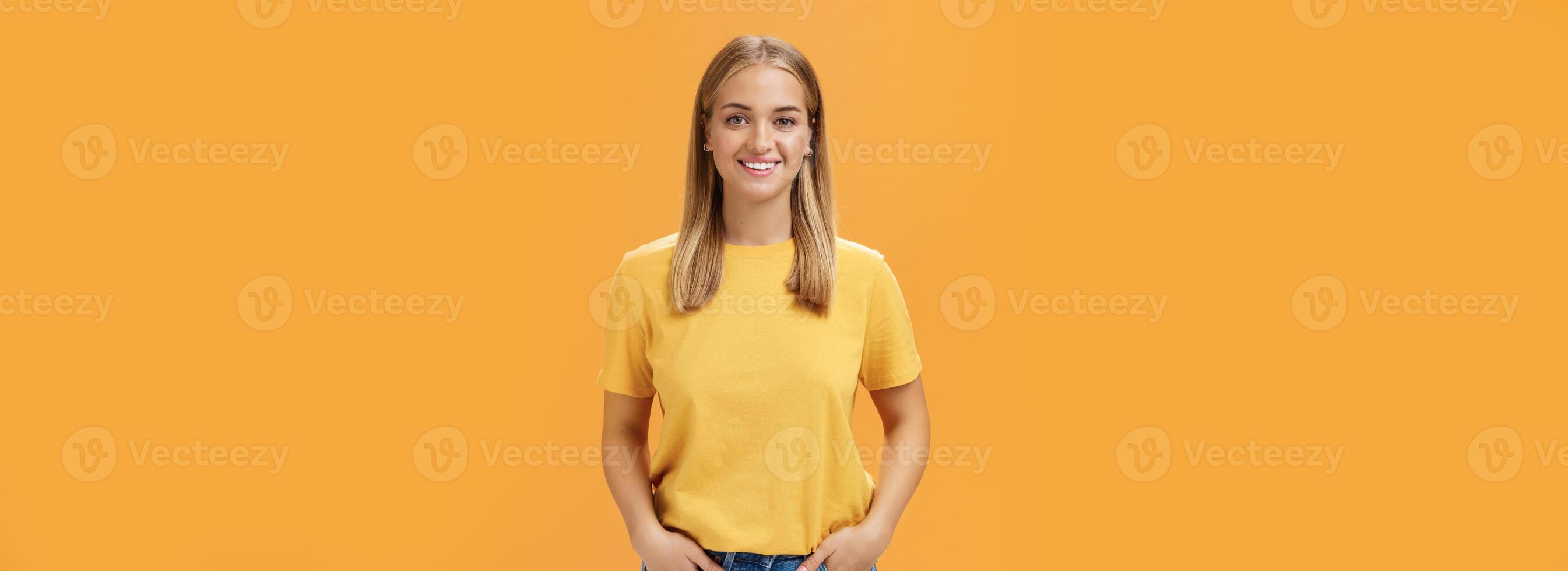 Cute chubby female with tanned skin and fair hair posing optimistic and joyful against orange background holding hands in pockets smiling broadly at camera charismatic and friendly-looking photo