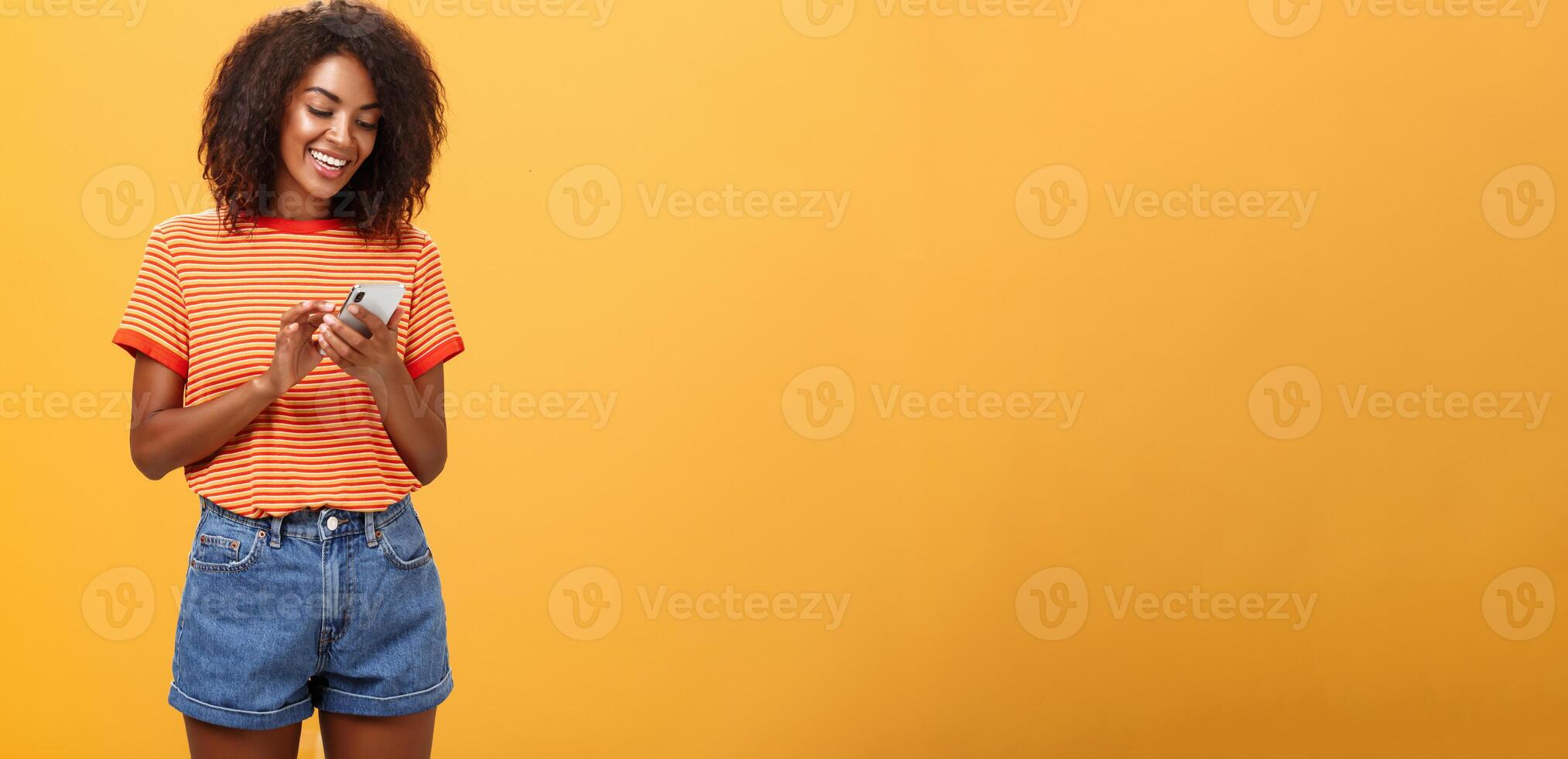 Stylish carefree girl texting friend come over standing pleased over orange wall in stylish denim shorts typing message or scrolling news in internet via smartphone gazing at device screen with smile photo