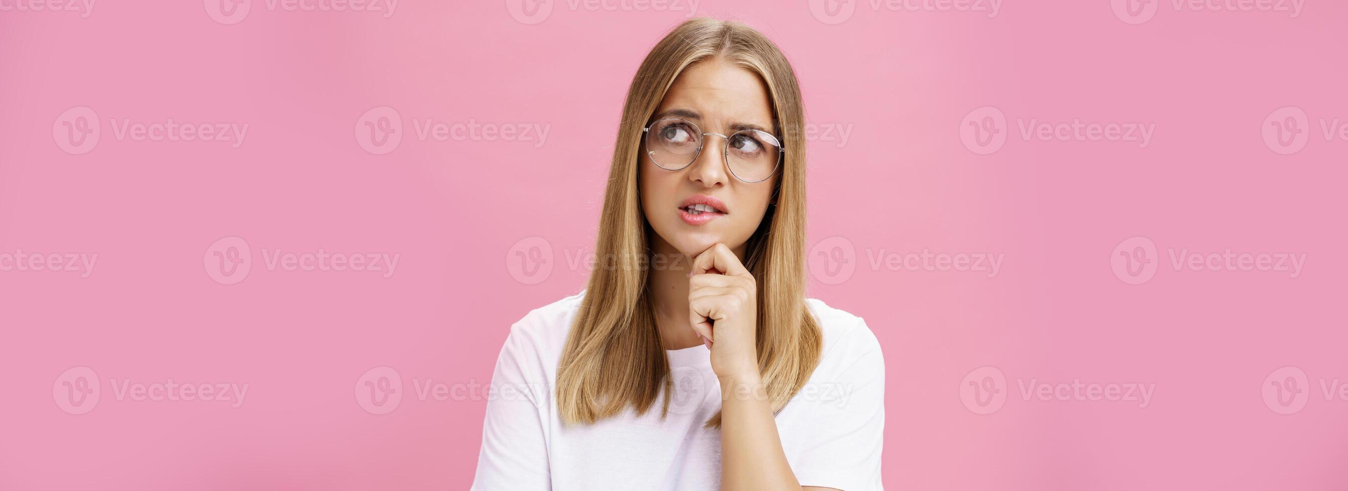 estudio Disparo de inseguro inteligente nerd mujer en lentes y blanco camiseta en pie preocupado y preocupado conmovedor barbilla mirando molesto a Superior Derecha esquina vacilando, pensando en contra rosado pared foto