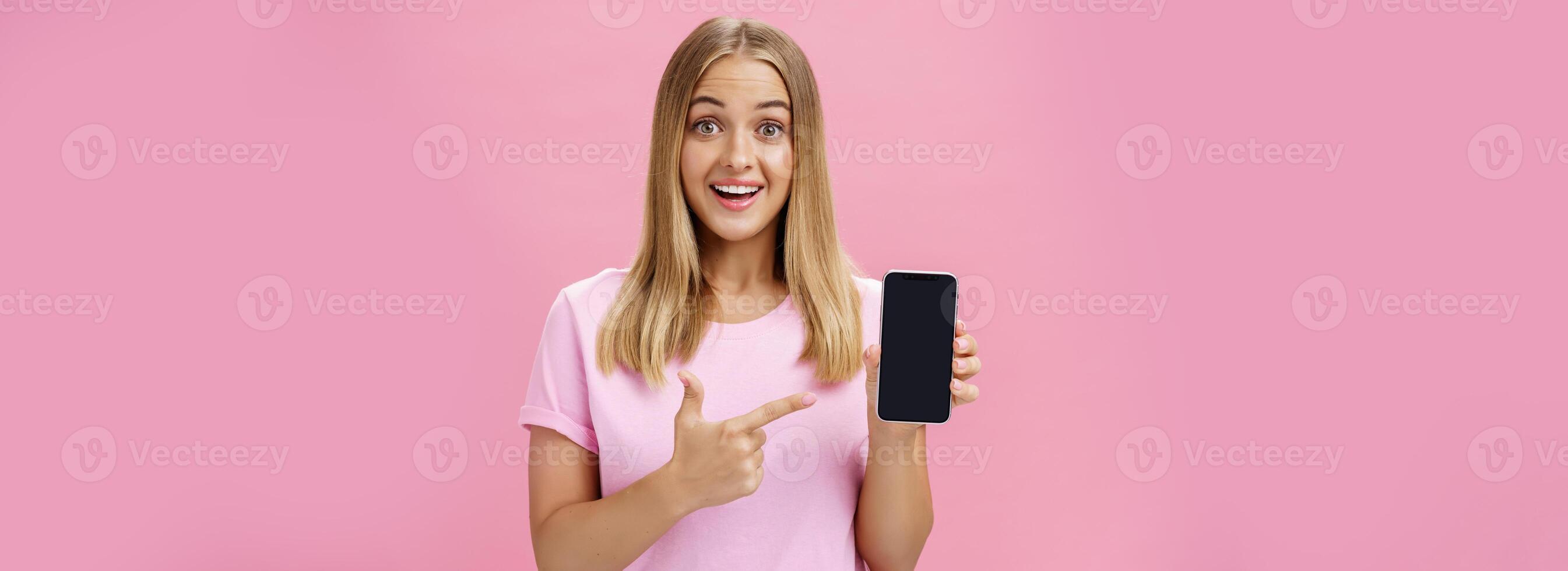 Cheerful attractive and pleasant woman promoting cool app or smartphone holding cellphone and pointing at device screen smiling amused and impressed standing over pink background photo