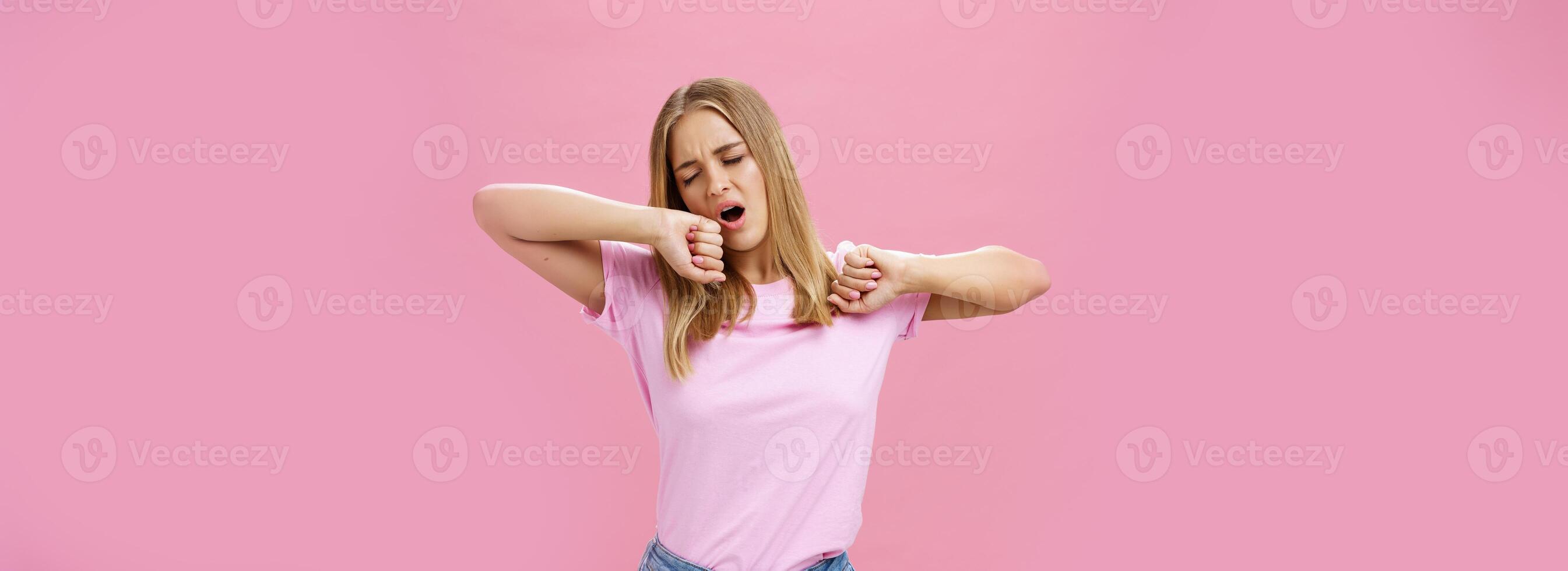 Too tired to work today. Lazy and exhausted attractive young female student doing homework all night yawning with closed eyes while stretching hands from after tiresome project over pink wall photo