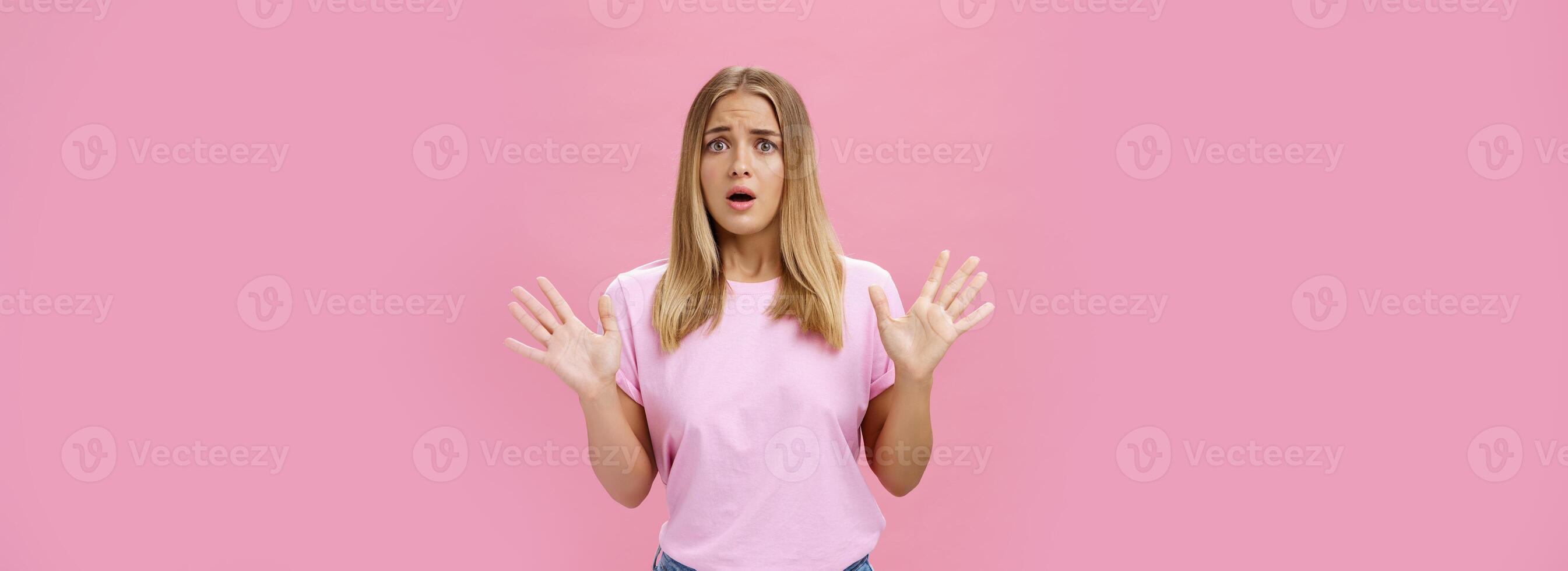 Woman looking nervous explaining with panicking gestures she not involved frowning opening mouth and gasping feeling concerned and worried waving hands over chest posing against pink wall photo