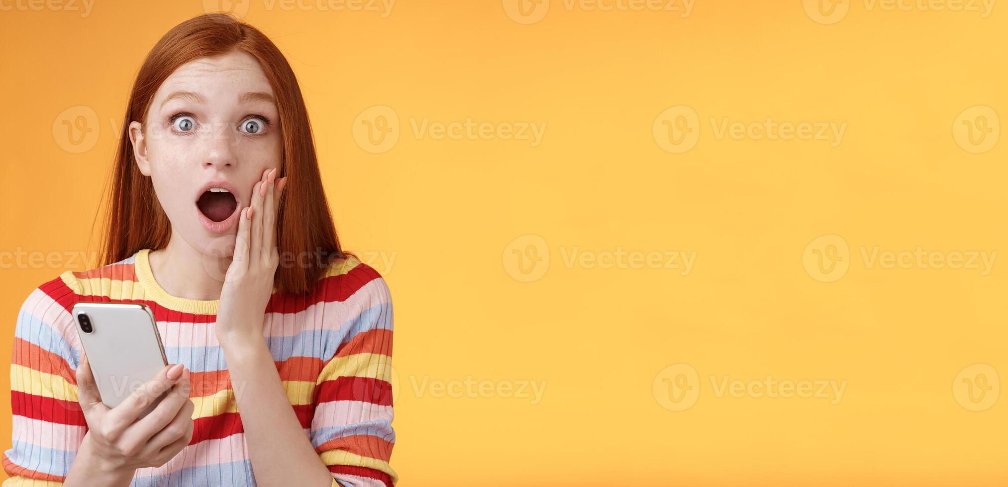 Concerned shocked emotive redhead girl find out stunning news look camera drop jaw gasping impressed touch cheek hold smartphone finish reading impressive disturbing story, orange background photo