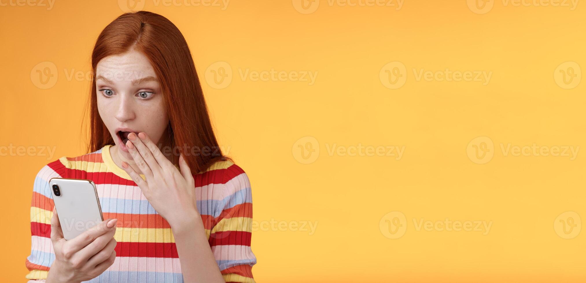 Amazed speechless young teenage redhead girl student gasping drop jaw say omg wow cover opened mouth palm look shocked surprised smartphone display reading fresh gossips orange background photo