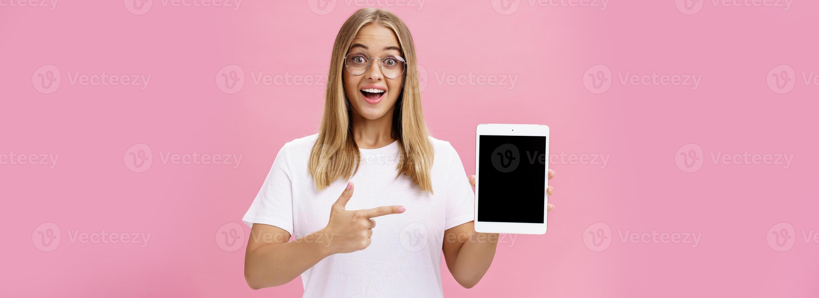 Girl suggests buy digital tablet for university and forget paper books. Excited happy and delighted young female student in white t-shirt and glasses pointing at device screen recommending gadget photo