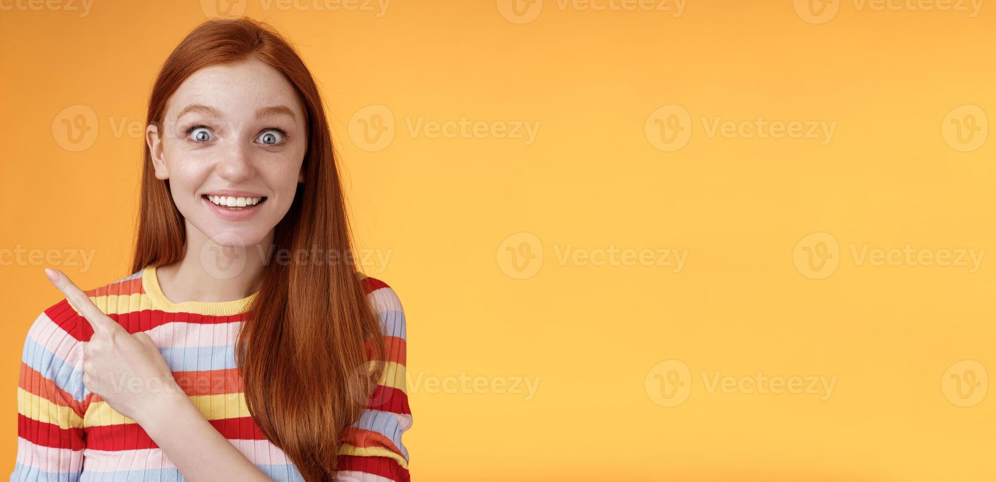 Excited thrilled good-looking young silly redhead girl surprised pop eyes camera gasping impressed pointing upper left corner see super star grinning excitement delight, orange background photo