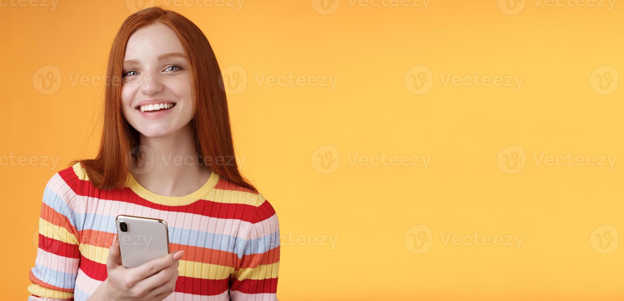 simpático entusiasta joven pelirrojo niña azul ojos utilizando teléfono inteligente giro cámara responder sonriente en general narración quien expedido mensaje en pie Encantado naranja antecedentes mensajería, mensajes de texto novio foto
