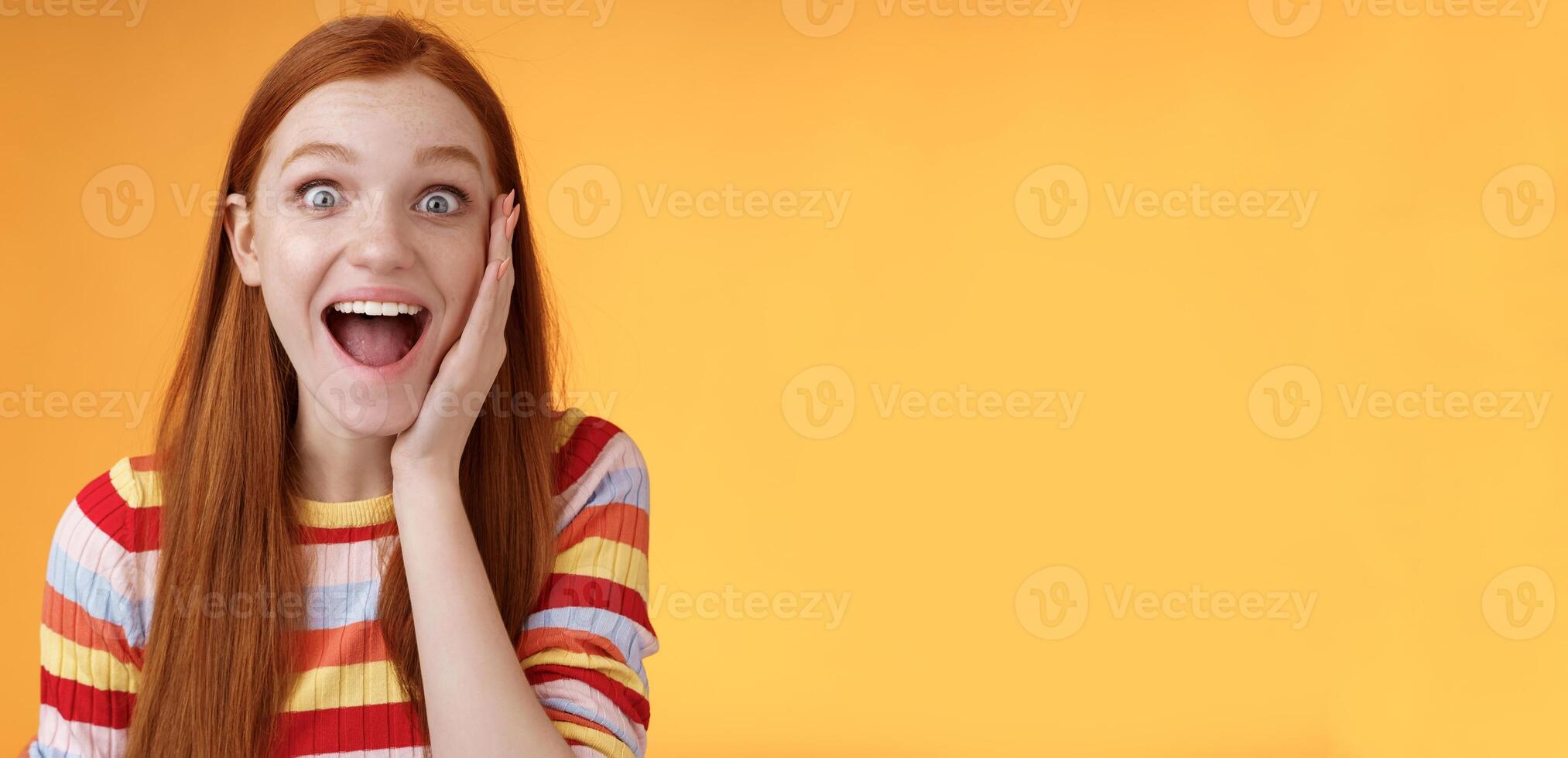 Amused happy smiling good-looking redhead woman screaming happily touch cheek astonished receive good perfect news triumphing feeling excited look thrilled camera, standing orange background photo