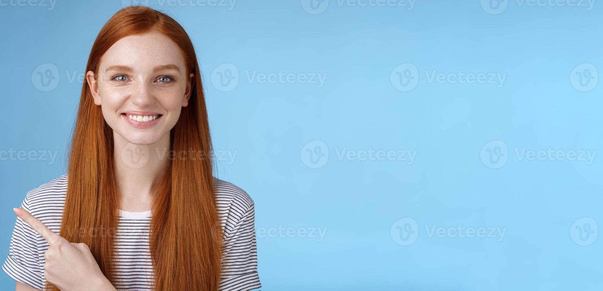 Pleasant charismatic helpful friendly-looking female redhead blue eyes pointing left index finger help make decision pick product store smiling broadly confident gaze camera asking trust her photo