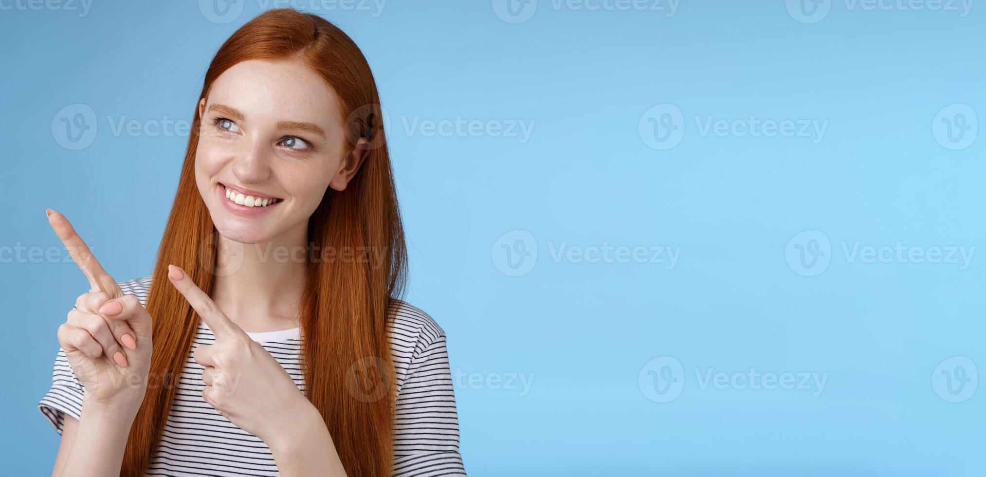Attractive redhead female model pure clean skin ginger long hair pointing looking left intrigued smiling satisfied indicating interesting advertisement intrigued using new product, blue background photo