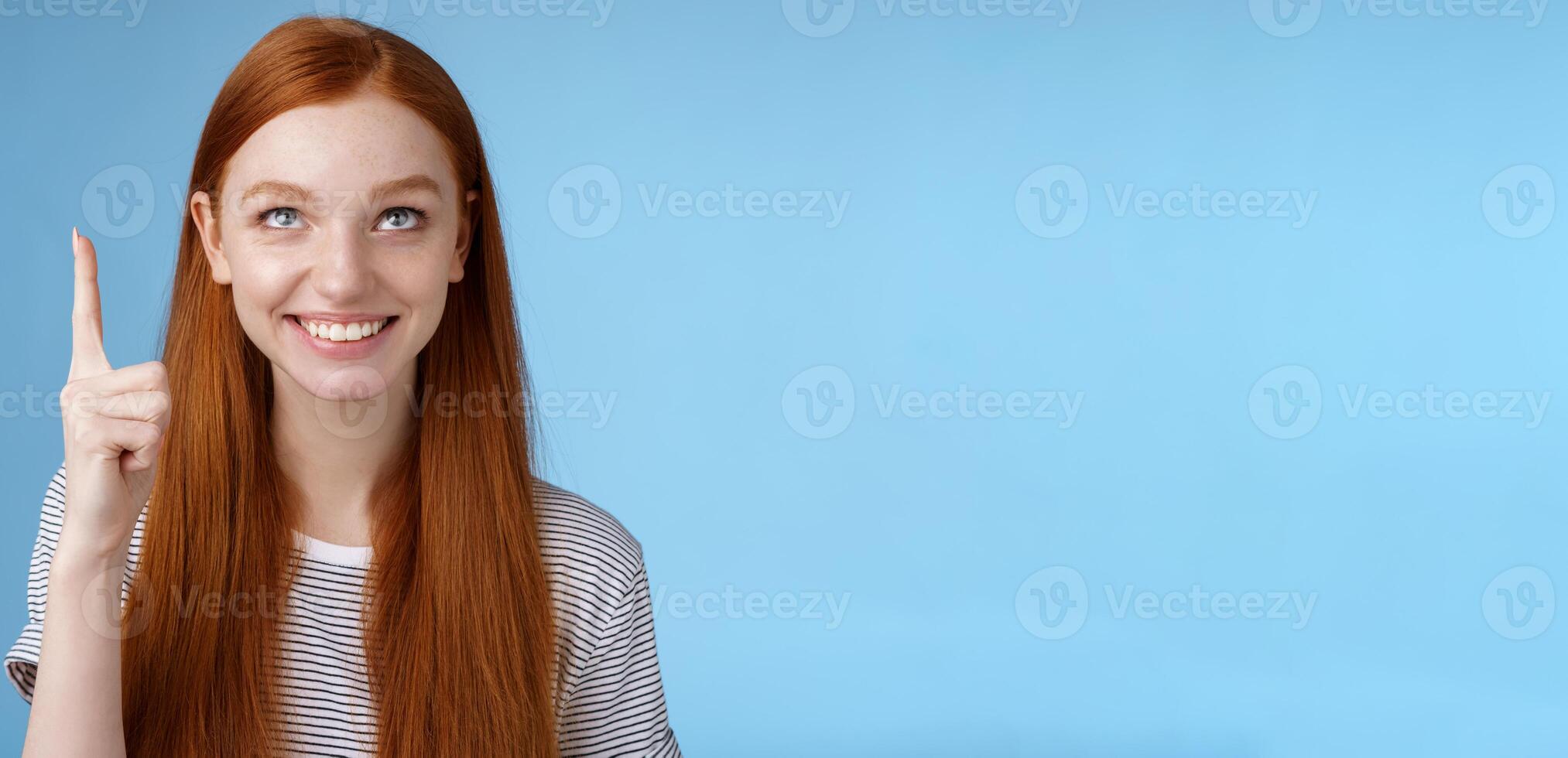 Intrigued lively happy smiling redhead european girl 20s look pointing up index fingers amused check out interesting sale holiday promo offer standing intrigued thrilled blue background photo