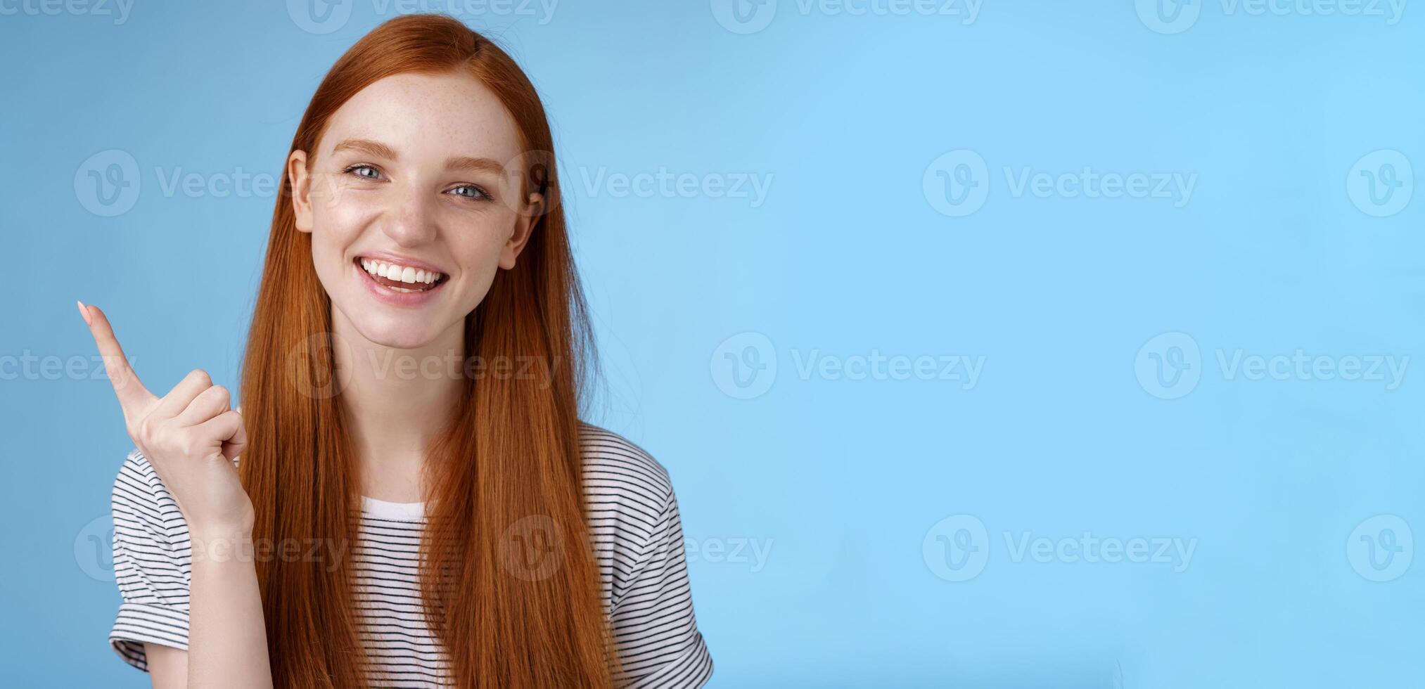 Happy charismatic redhead laughing young girl having fun looking carefree talking discussing new product sale pointing up index finger showing copy space advertisement, standing blue background photo