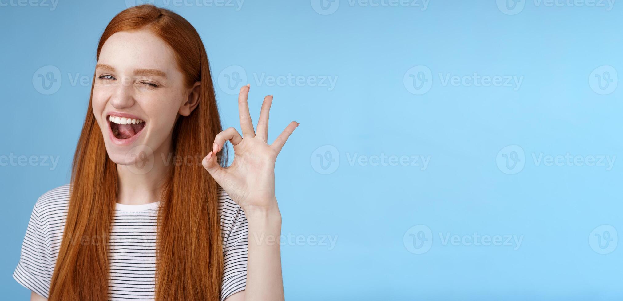 Sassy attractive redhead girl winking mysteriously smiling broadly give approval sign show okay ok excellent gesture satisfied good choice agree great decision, standing delighted blue background photo