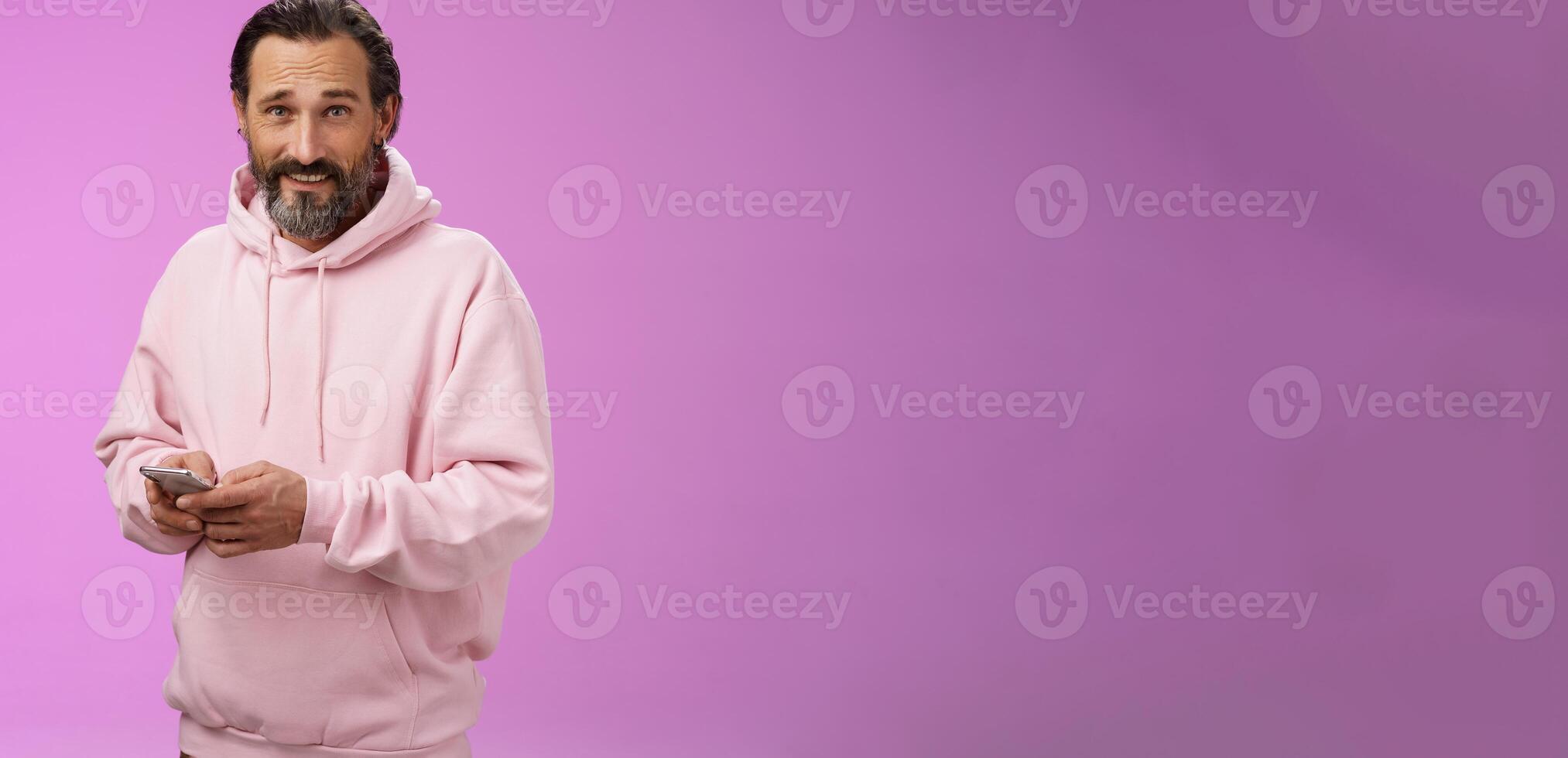 Stylish handsome adult bearded guy 40s in trendy pink hoodie holding smartphone texting look camera unbothered cool confident distracted playing awesome new app game, purple background photo