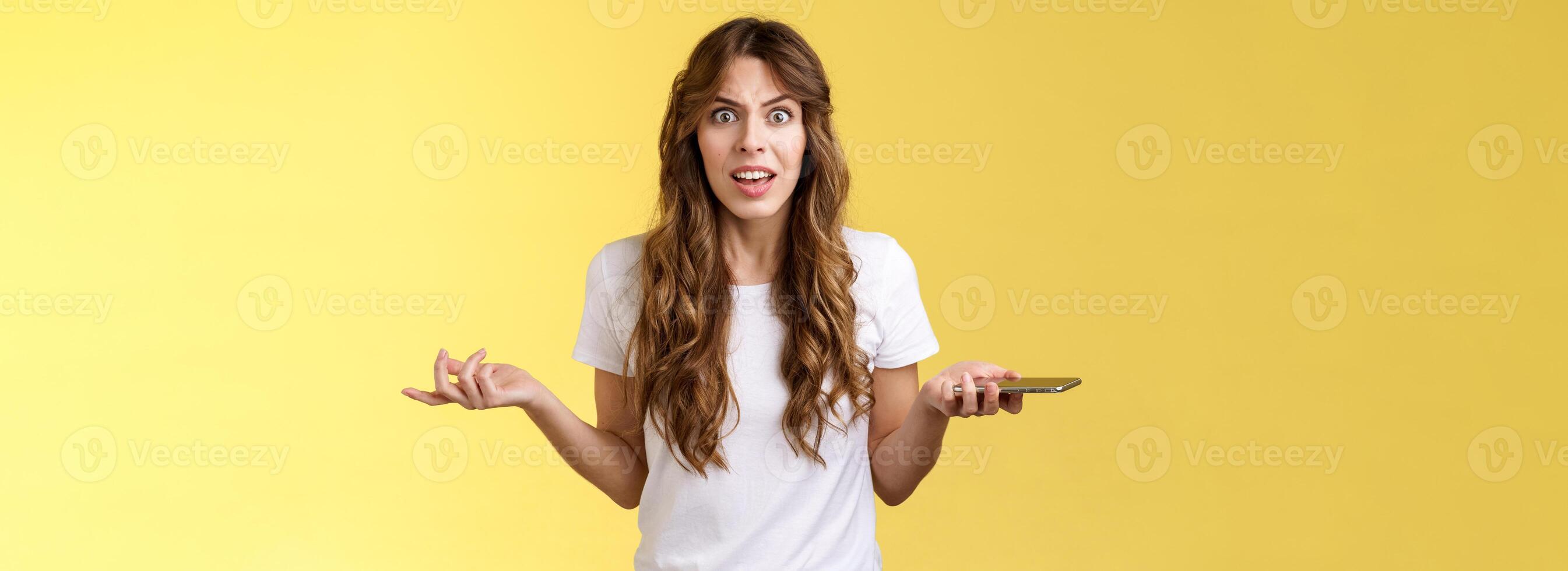 Pissed frustrated bothered curly-haired caucasian girl stare camera puzzled complain hold smartphone shrugging spread hands sideways dismay arguing after intense serious phonecall yellow background photo
