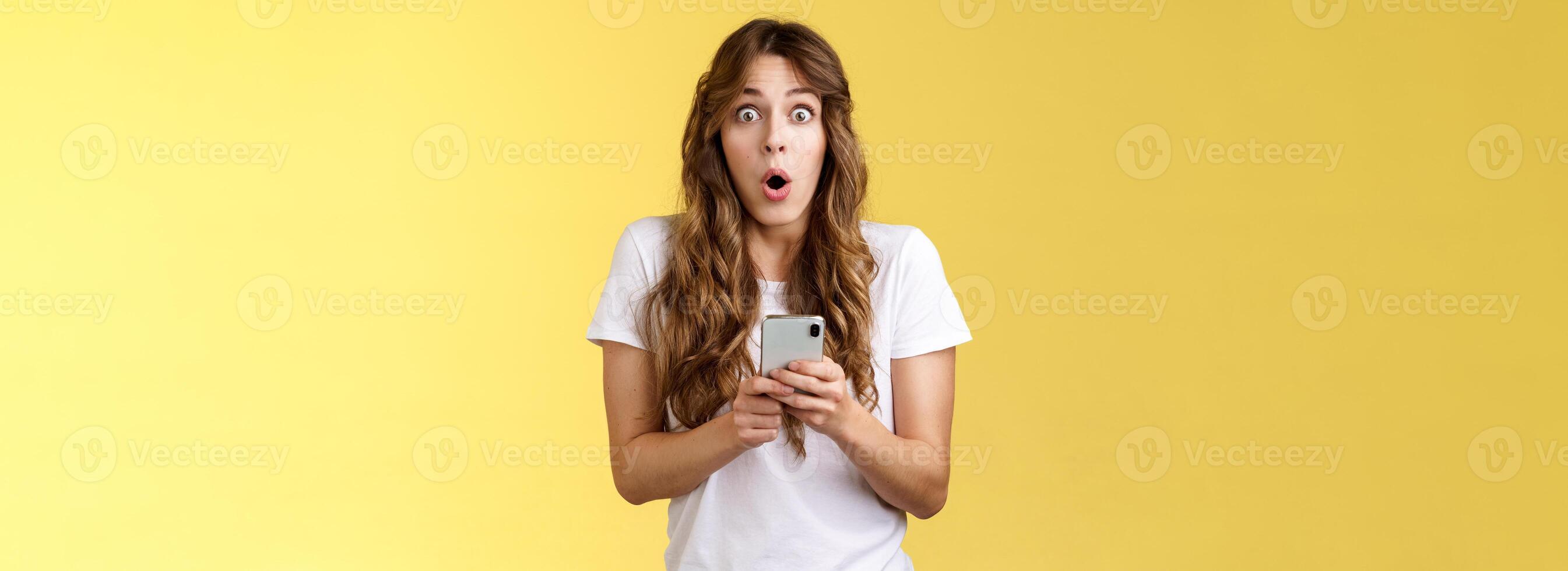 Shocked impressed speechless girl say wow curly hairstyle stare camera astonished hold smartphone reacting stunning perfect news receive awesome message stand yellow background photo