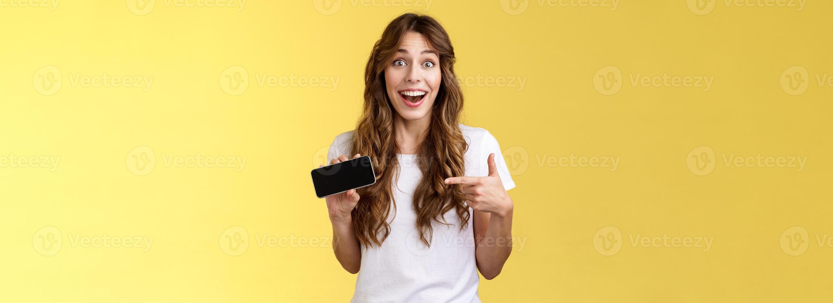Cheerful surprised cute lucky girl beat best score awesome game showing smartphone display pointing index finger horizontal screen mobile phone introduce cool app smiling broadly yellow background photo
