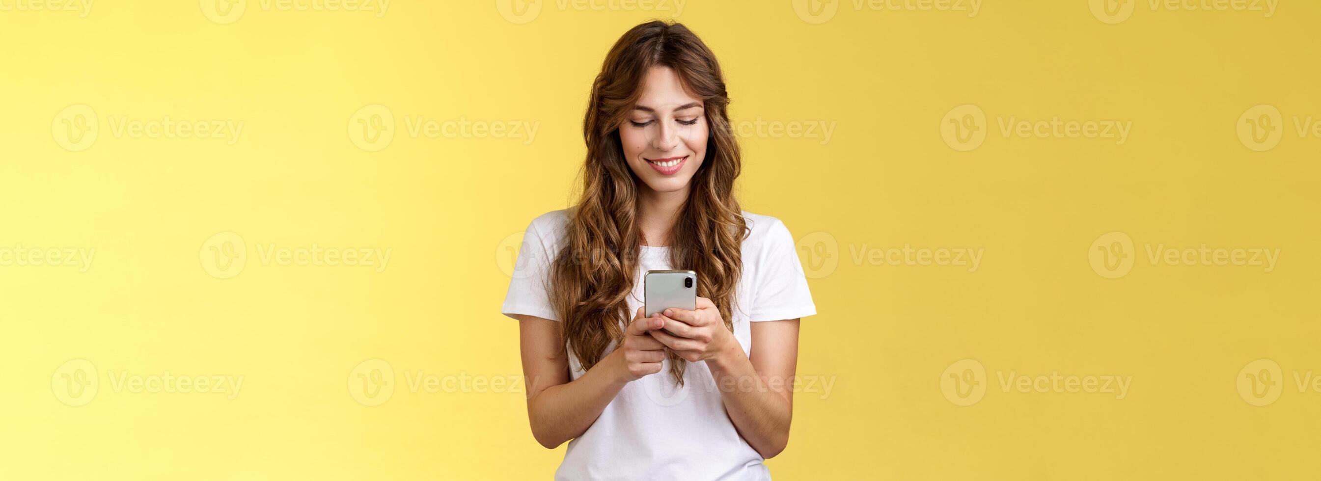 Lovely feminine tender caucasian girl white t-shirt jeans hold smartphone texting messaging girlfriend smiling delighted look tenderly lovely smile mobile phone screen yellow background photo