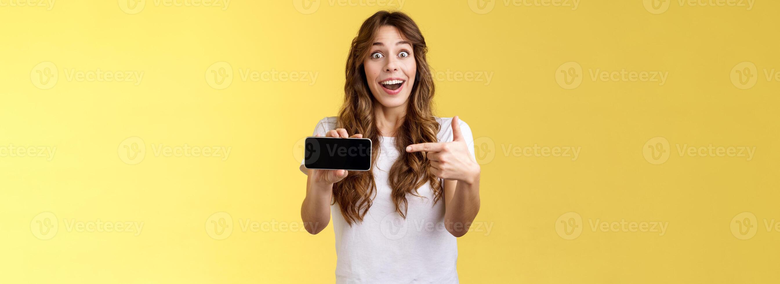 Impressed upbeat happy lucky girl curly long hairstyle open mouth admiration joy like awesome new app show smartphone screen horizontal phone display stand yellow background amazed photo