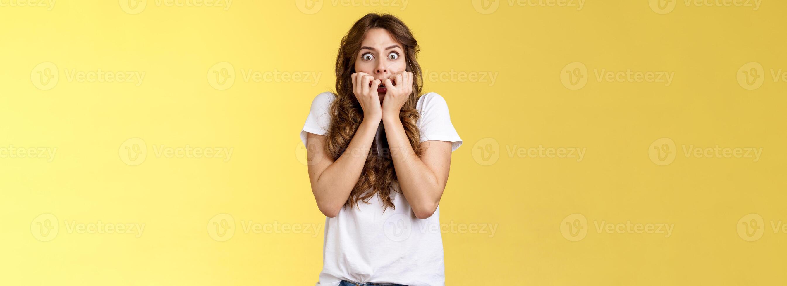 Shocked speechless scared stunned curly-haired caucasian woman gasping hold fingers mouth biting nails stare camera frightened express fear terrified scary movie stand yellow background photo