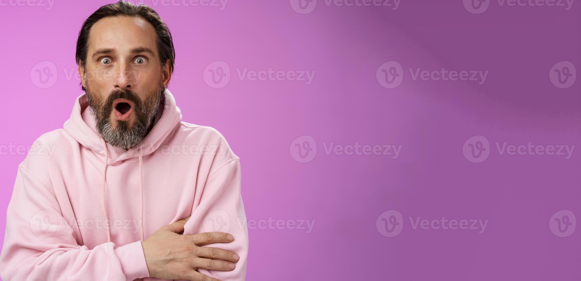 Shocked impressed gossiping caucasian bearded adult 40s man grey hair in pink hoodie gasping fascinated fold lips wow widen eyes astonished hearing interesting thrilling story, purple background photo