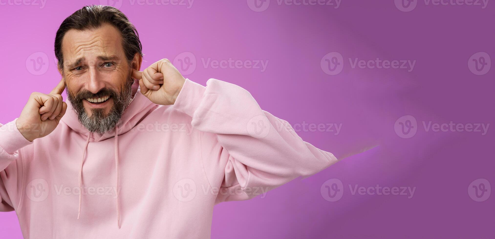 Displeased annoyed adult bearded man feel discomfort distracted loud awful noise frowning cringing hear bad musician play guitar put fingers ears grimacing asking turn volume off, purple background photo
