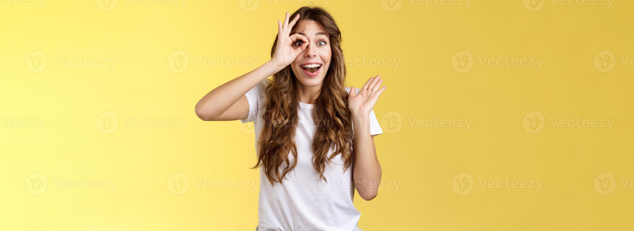 Oh hi I see you. Fascinated cute charming young girl waving palm say hello impressed look through okay ring gesture impressed finally fixed sight stand astonished amazed yellow background photo