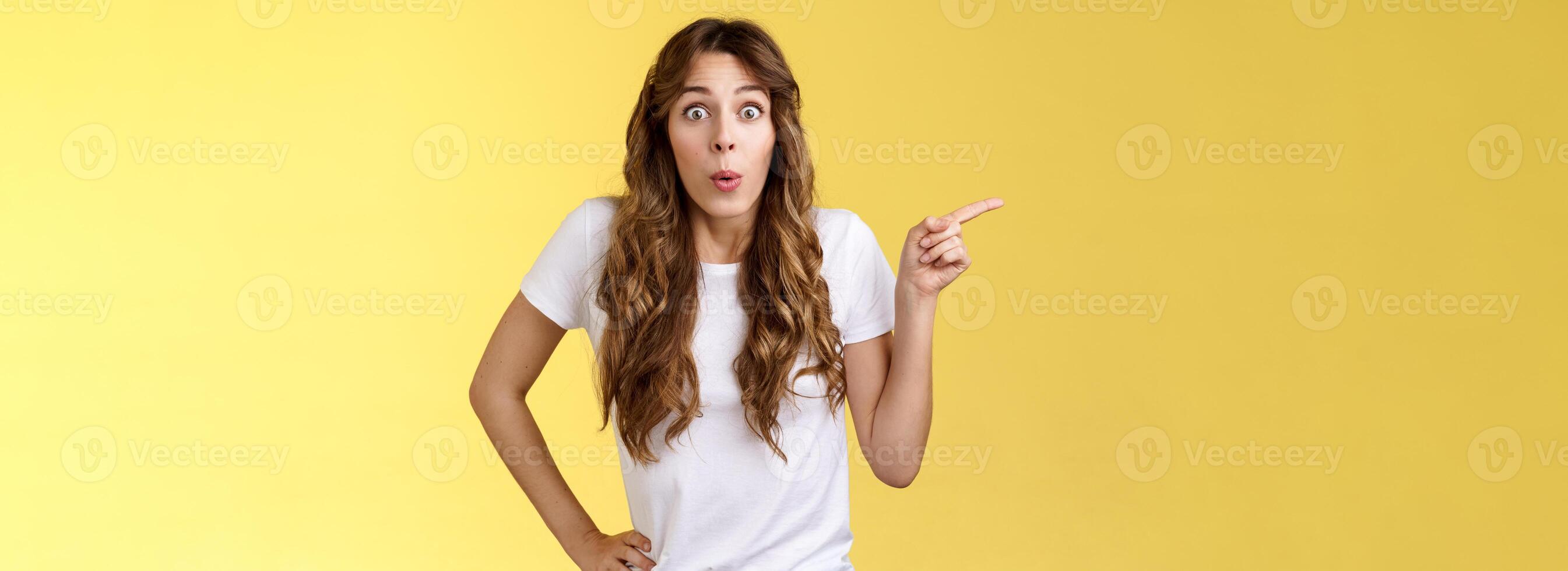 Wow incredible lets click. Impressed surprised pretty european woman long curly hairstyle folding lips intrigued stare astonished wondered awesome cool promo offer pointing left yellow background photo