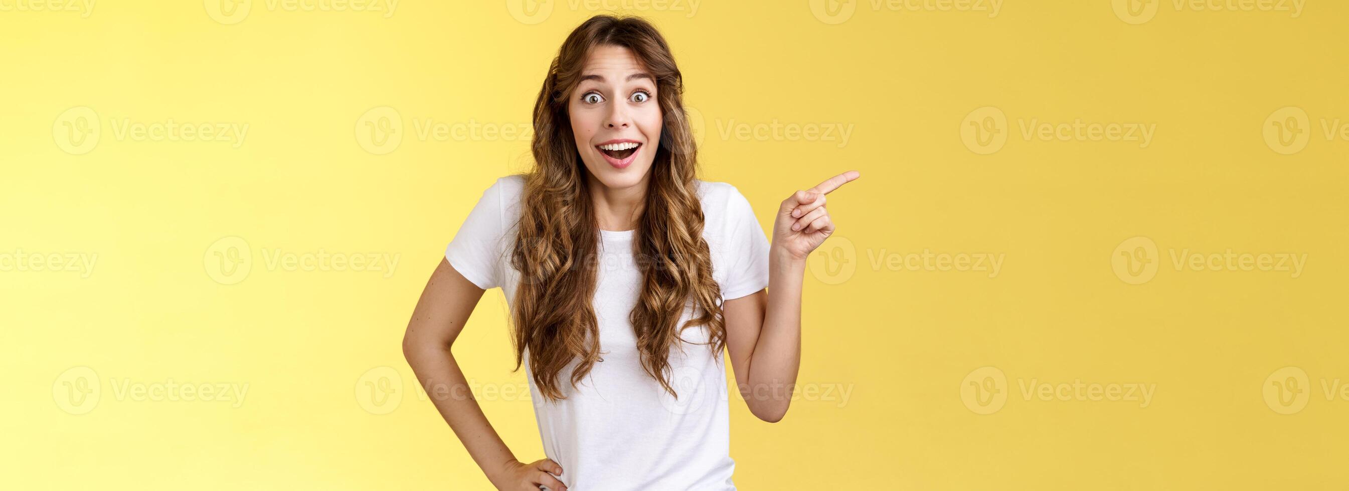 Surprised friendly sociable happy cute girlfriend long curly chestnut haircut smiling impressed gladly pointing left grinning camera toothy astonished expression discuss awesome performance photo