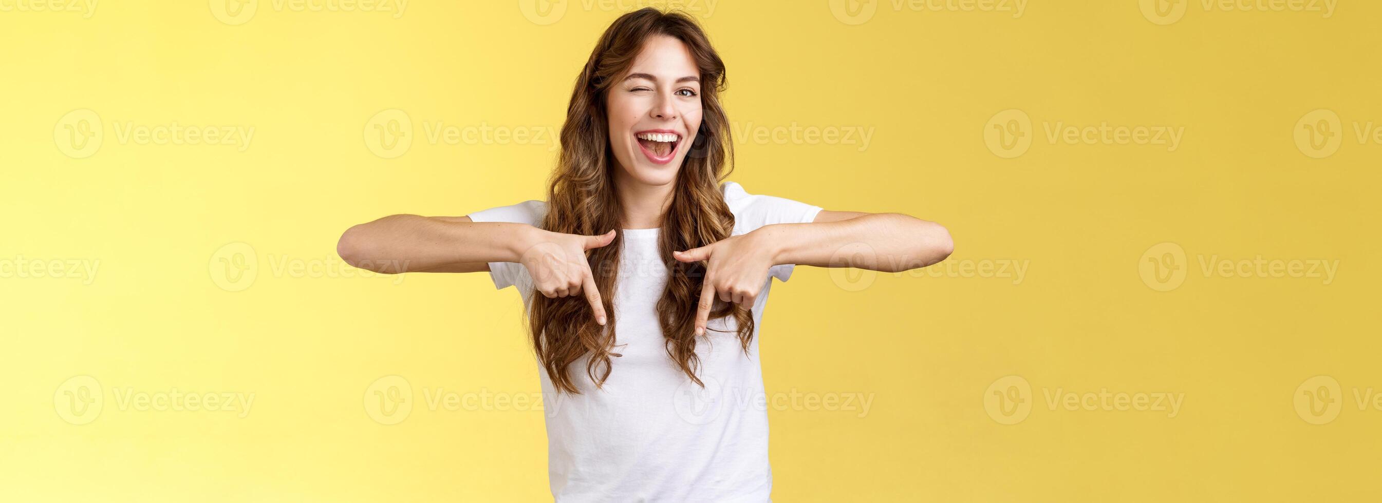 Hey visit my link would not regret. Lively cheeky enthusiastic woman curly long haircut wink sassy smiling broadly show you perfect copy space pointing down bottom promo yellow background photo