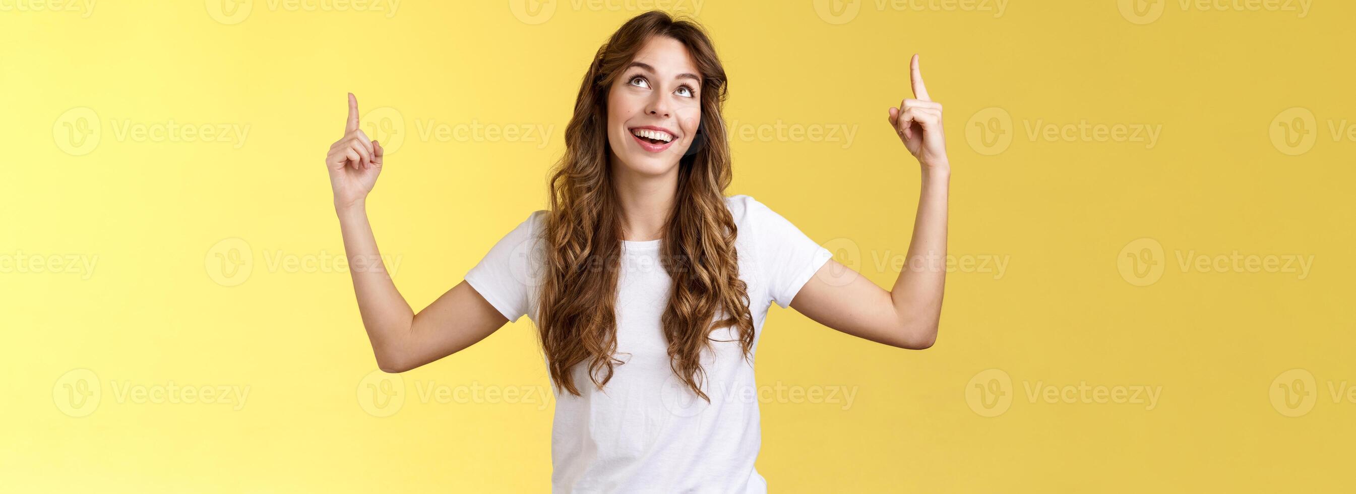 Happy admiring cute european curly-haired girl long haircut look pointing up impressed amused smiling broadly satisfactory delighted stand white t-shirt yellow background joyfully react good promo photo